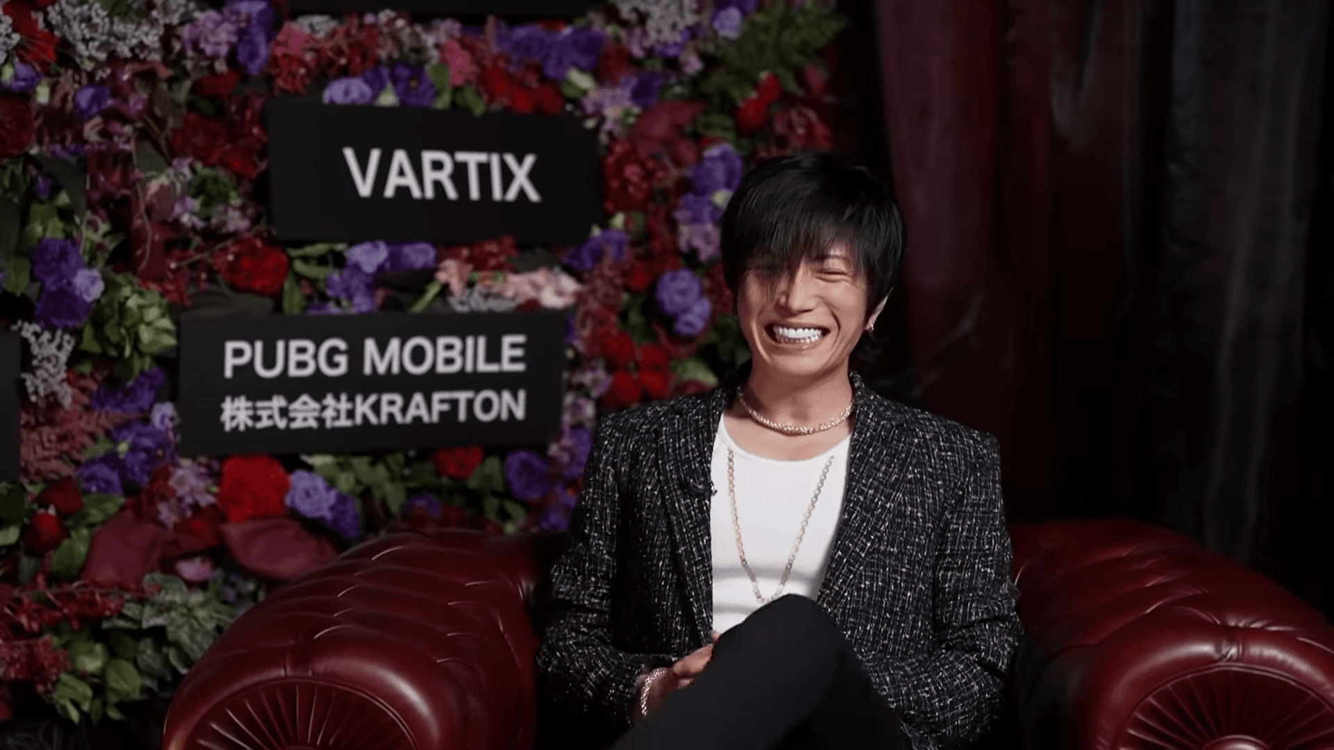 紫と赤の花の壁の前で笑う GACKT