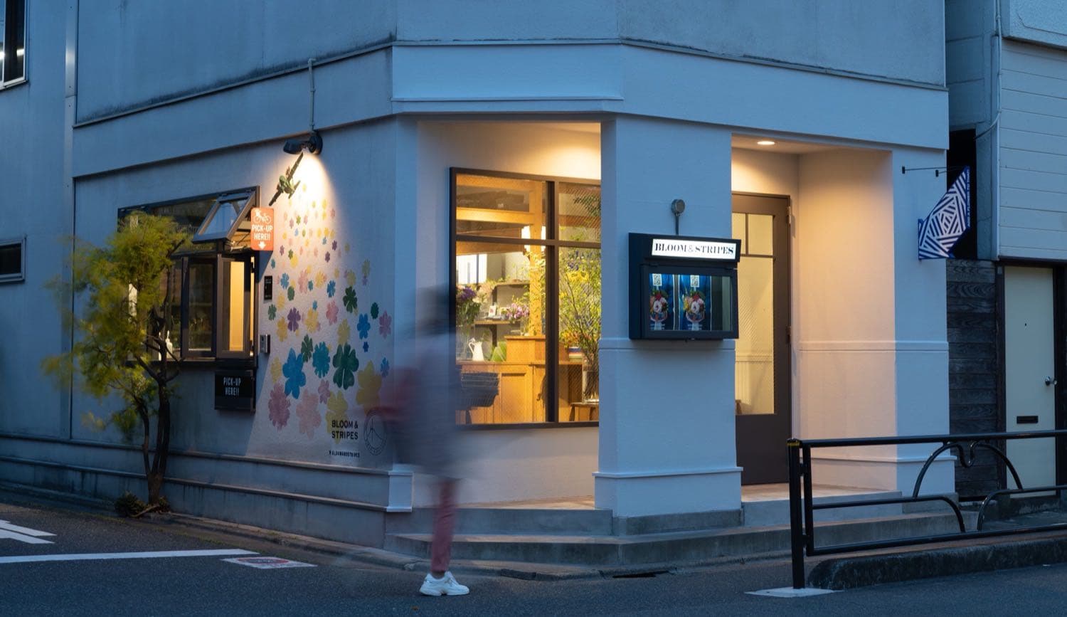 夕暮れの角地に建つ自由が丘店の外観。花のイラストを描いた白い壁と、明かりのともったガラス張りの店内